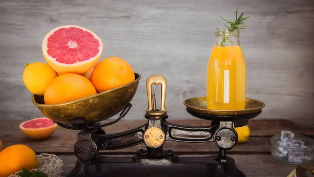 A photo representing grapefruit vs orange comparison showing whole grapefruits on one side and a bottle of orange juice in a vintage scale.